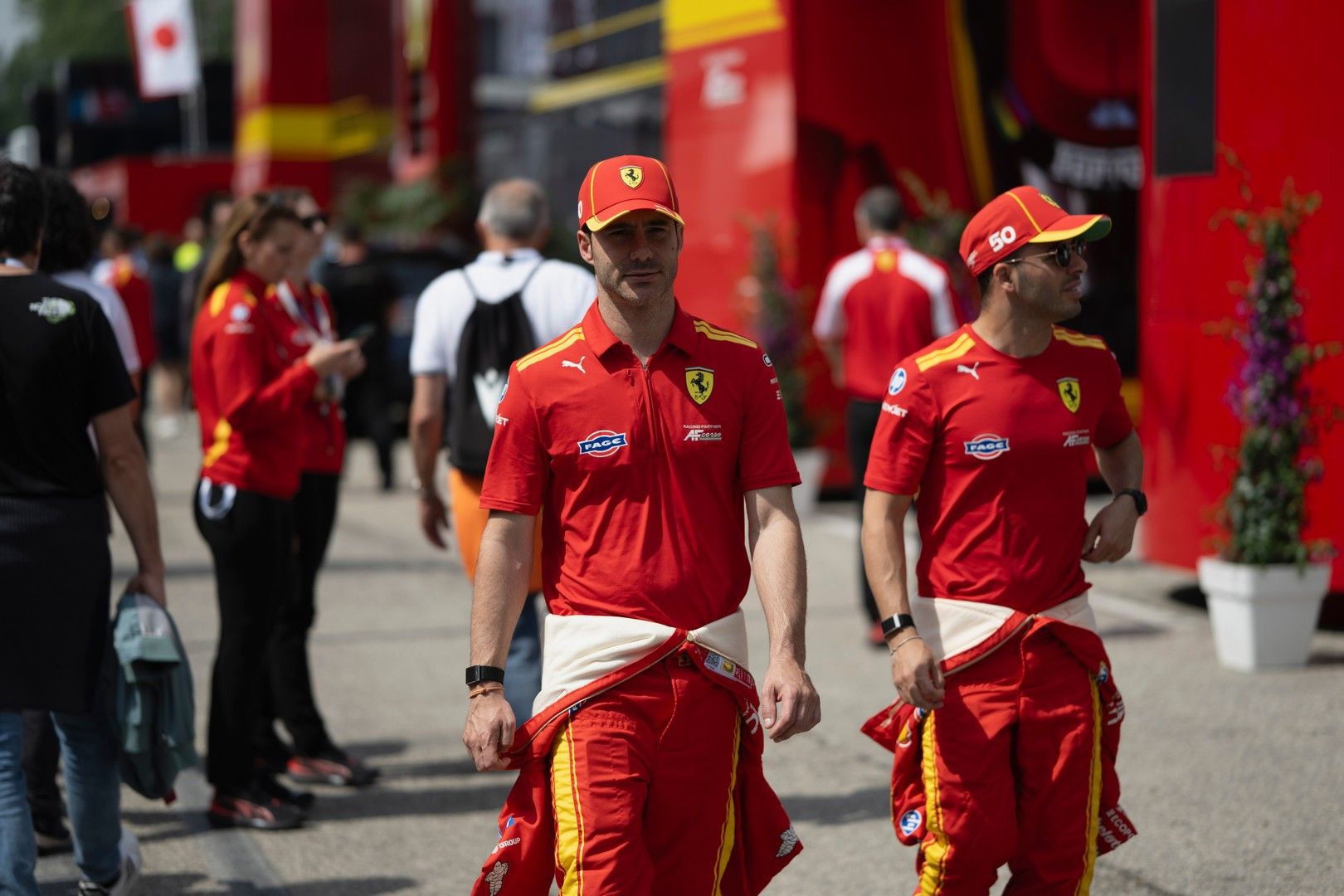 Two men in red Ferrari team shirts, caps, and racing suits tied at their wawaists walk outdoors.