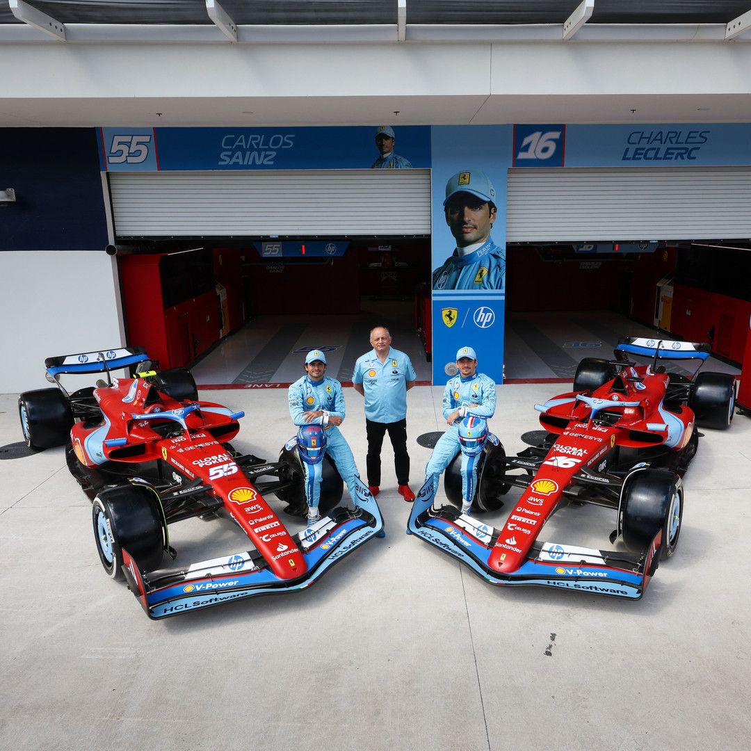 Ferrari F1 drivers Carlos Sainz and Charles Leclerc, in blue suits, on their red and blue F1 cars, with a team member in a pitlane.