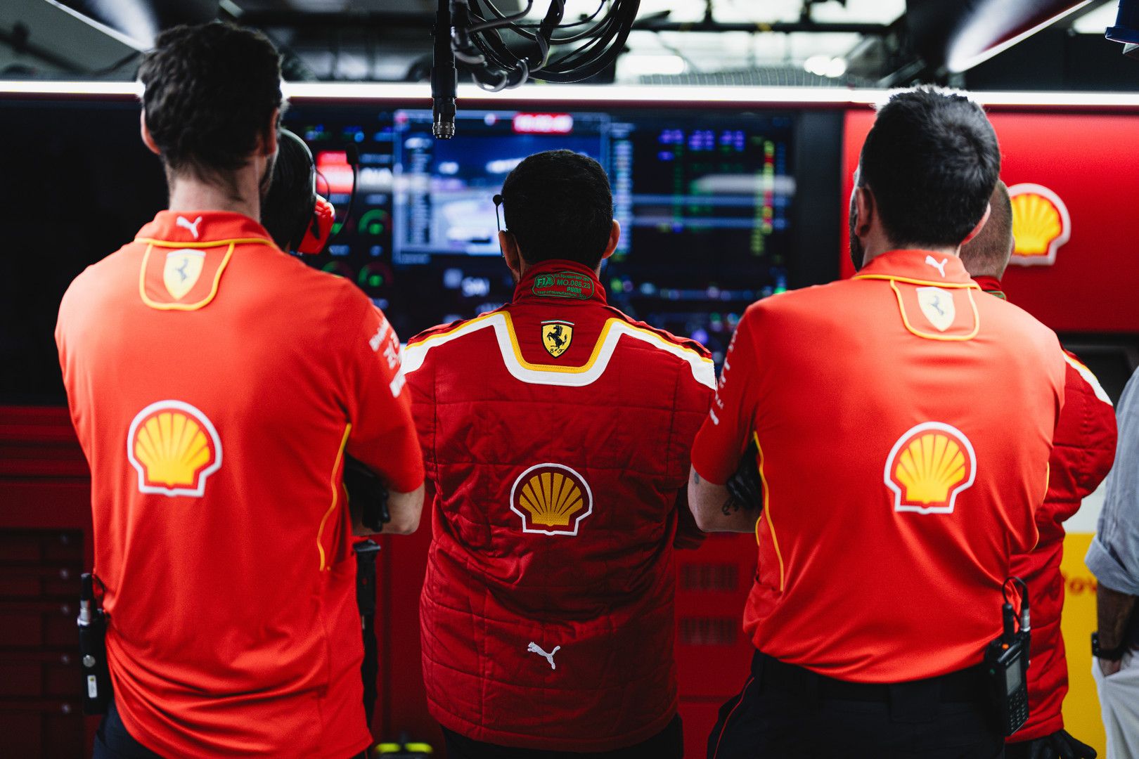 Three people in red Ferrari and Shell team uniforms, seen from behind, watching multiple computer screens displaying data.