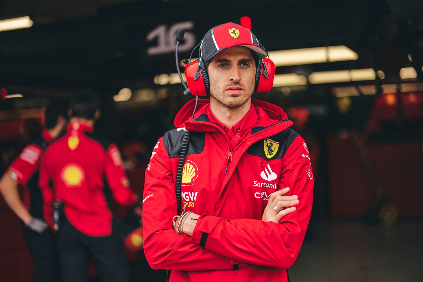 A man in a red Ferrari team jacket, cap, and red headphones stands with crossed arms in a garage.