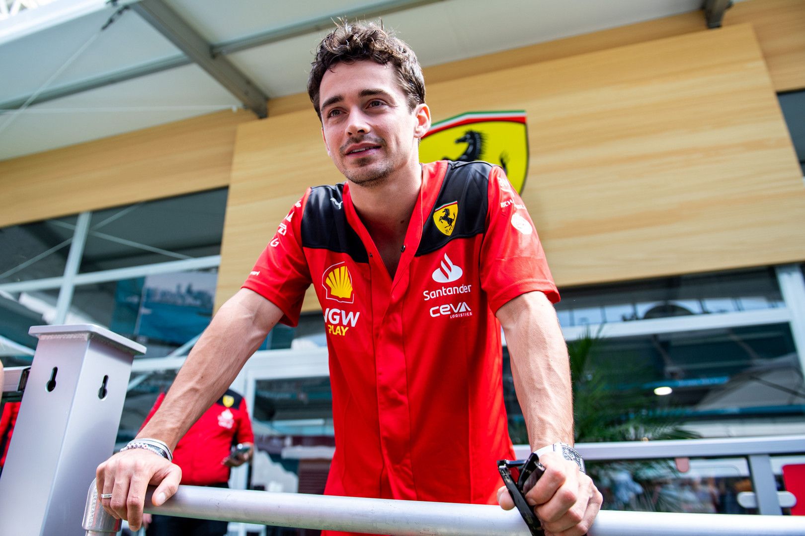 Charles Leclerc in a red Ferrari team shirt leans on a railing with the Ferrari logo in the background.