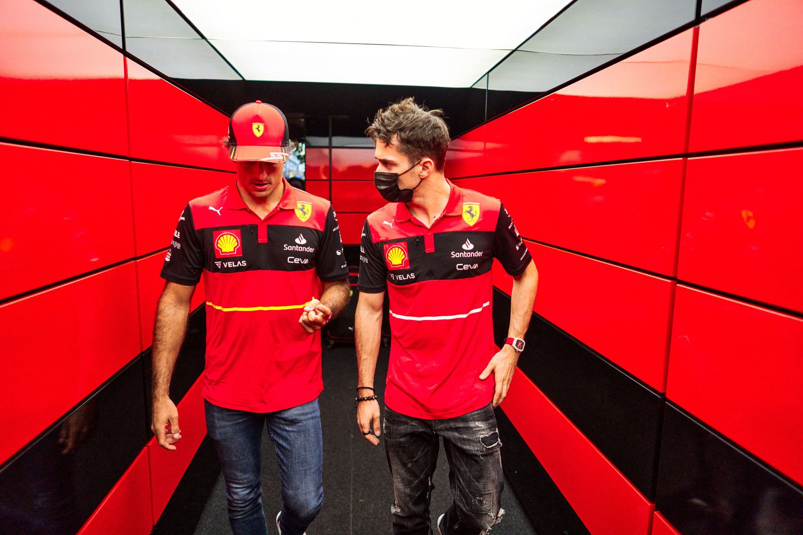 Two men wearing Ferrari team shirts walk down a red and black corridor.
