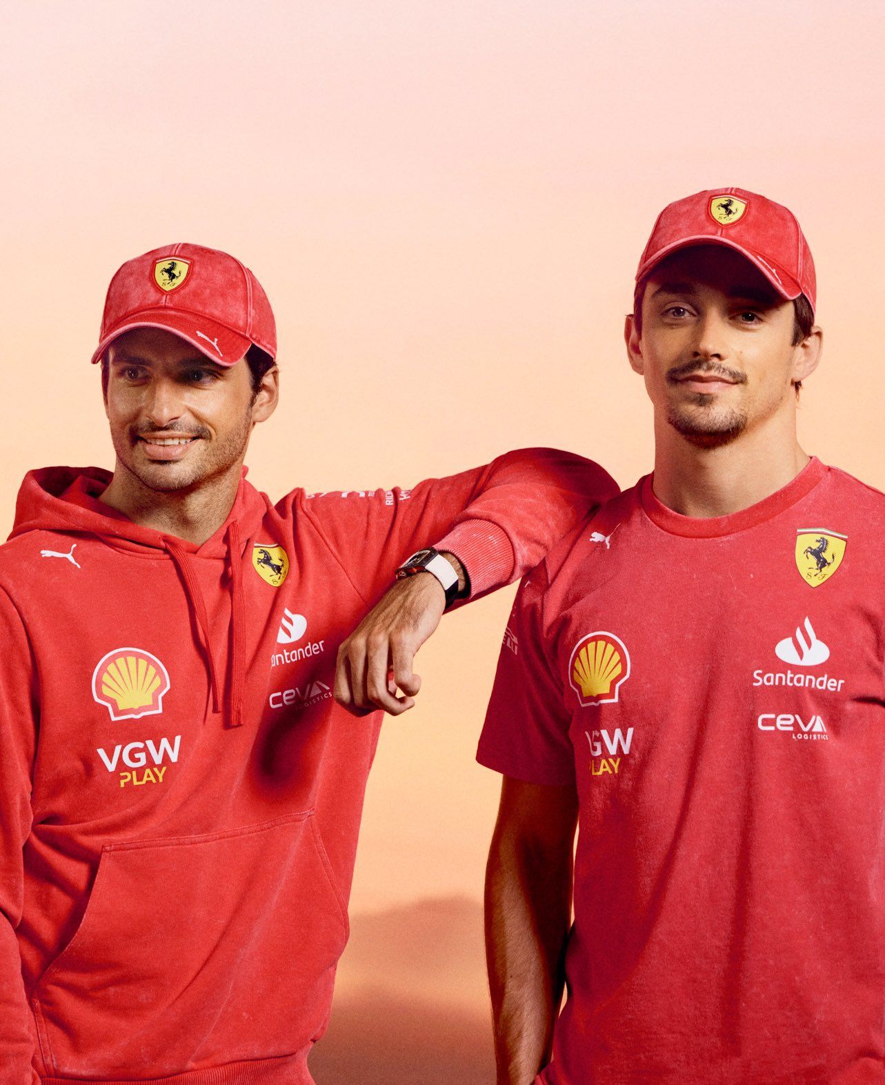 Ferrari F1 drivers Carlos Sainz and Charles Leclerc smiling in red team caps and apparel.