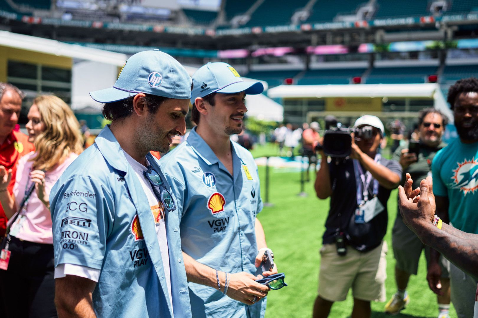 Two smiling Formula 1 drivers, Charles Leclerc and Carlos Sainz, in light blue team shirts and caps, walk across a sunny football stadium field.