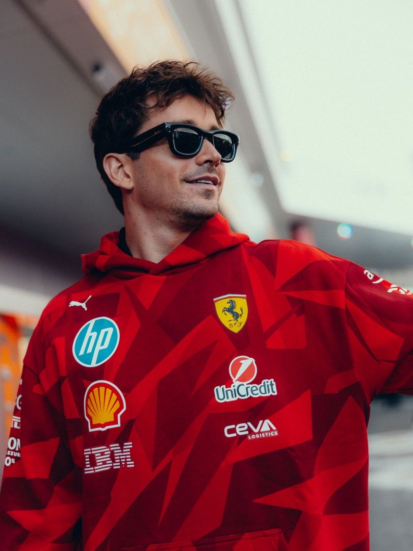 Charles Leclerc wearing sunglasses and a red Ferrari team hoodie covered in sponsor logos.