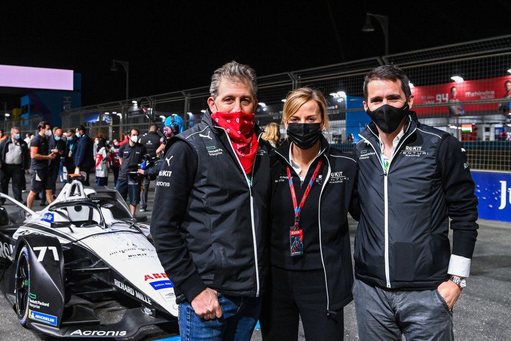 Three team members, two men and a woman, wearing jackets and face masks, stand on a race track at night next to a Formula E car with number 71.