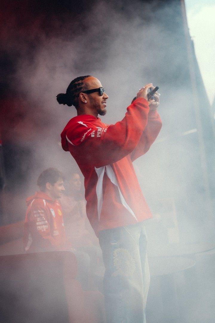 Lewis Hamilton in a red hoodie and sunglasses, holding a phone up to film amidst smoke.