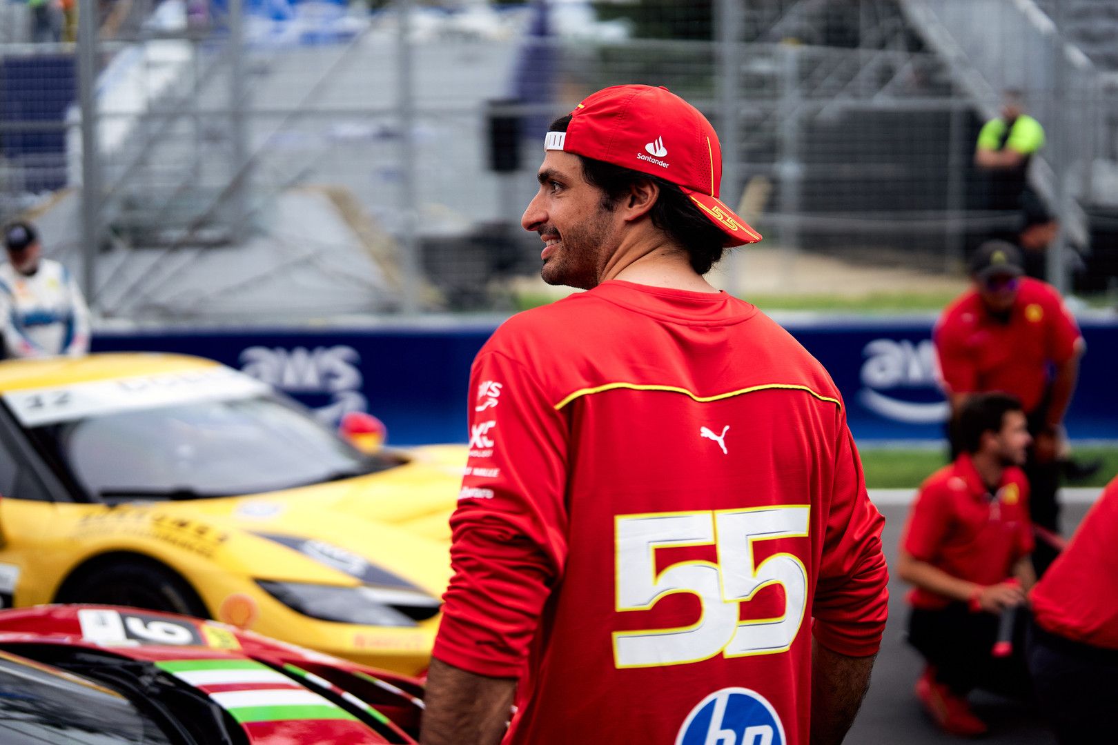 A smiling man in a red #55 shirt looks sideways on a race track with yellow and red race cars.