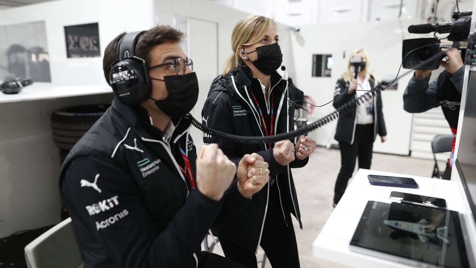 Two masked race crew members with headsets clench their fists in a control room; a tablet on the table shows a race car.