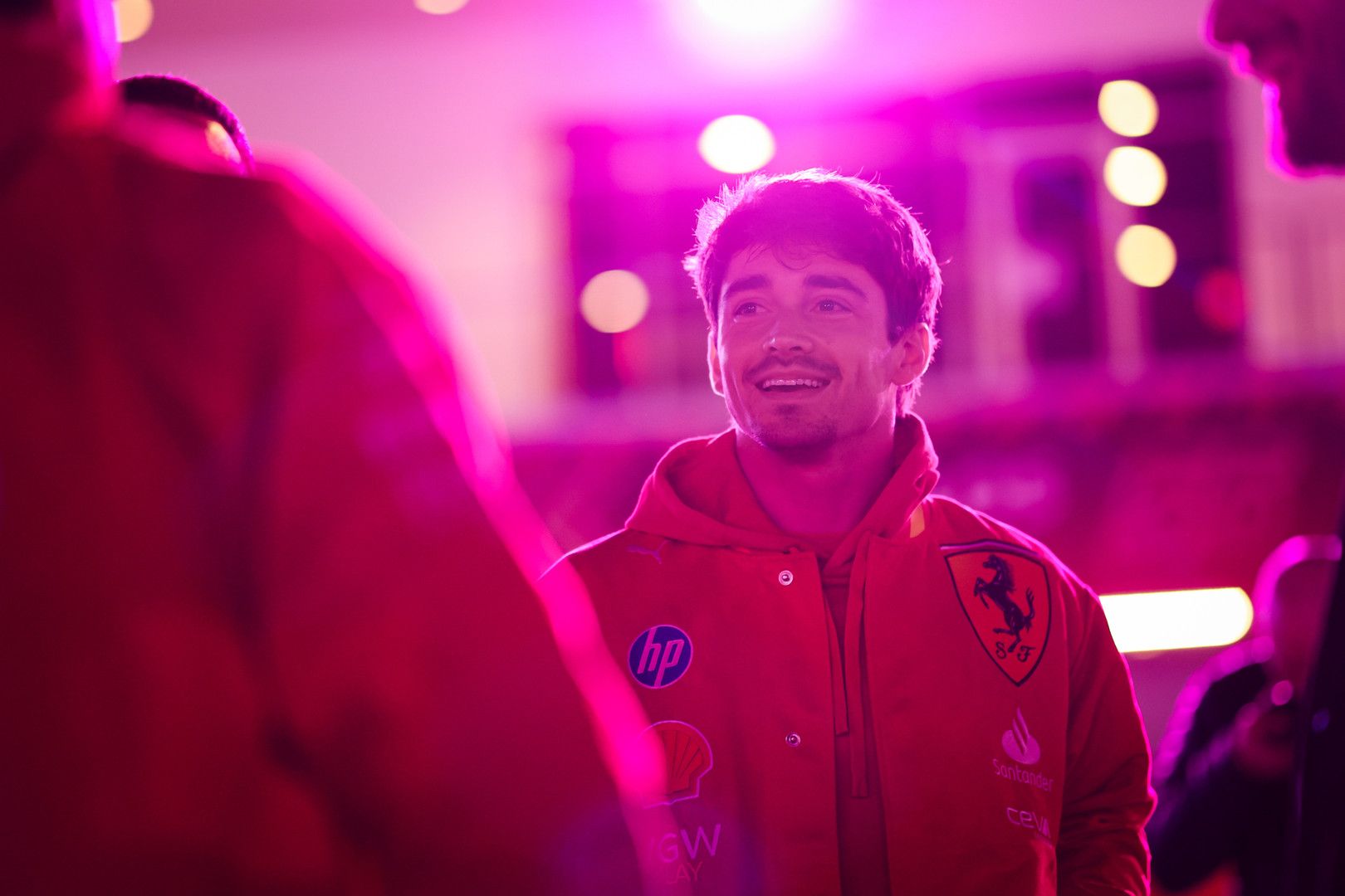 Charles Leclerc smiles in a red Ferrari jacket under magenta lighting.