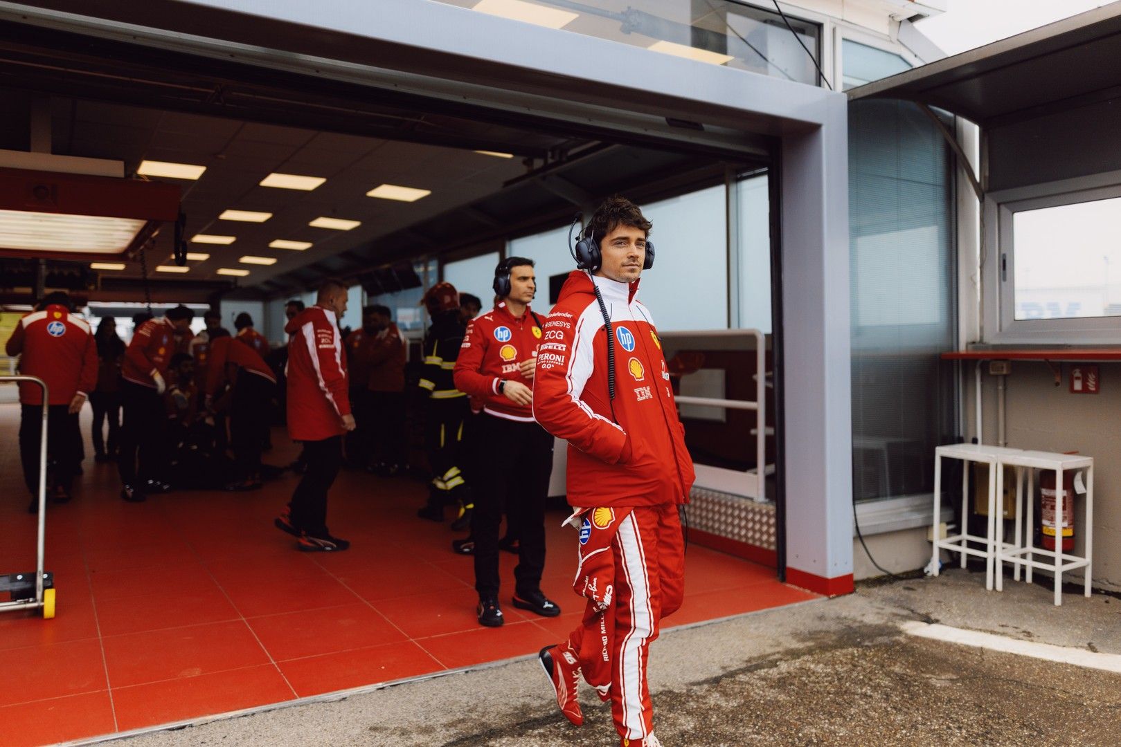 Charles Leclerc in a red Ferrari racing suit and headphones walks out of a pit garage.