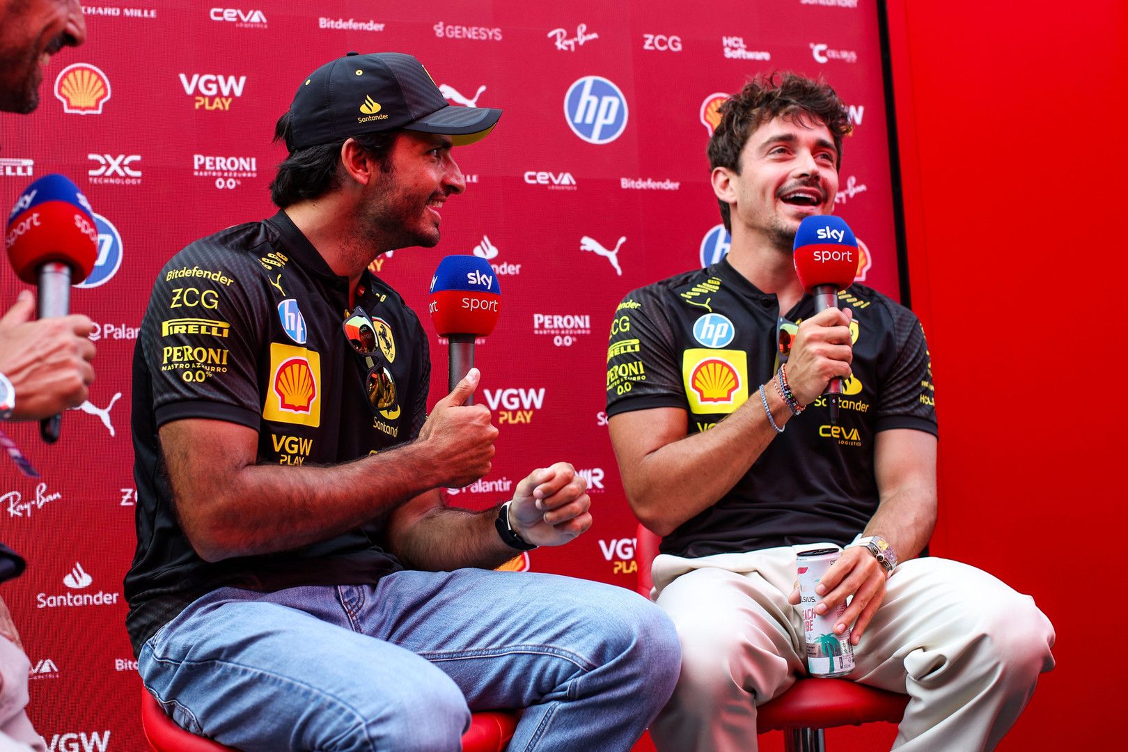 Carlos Sainz Jr. and Charles Leclerc smile, holding Sky Sports microphones at a press conference.
