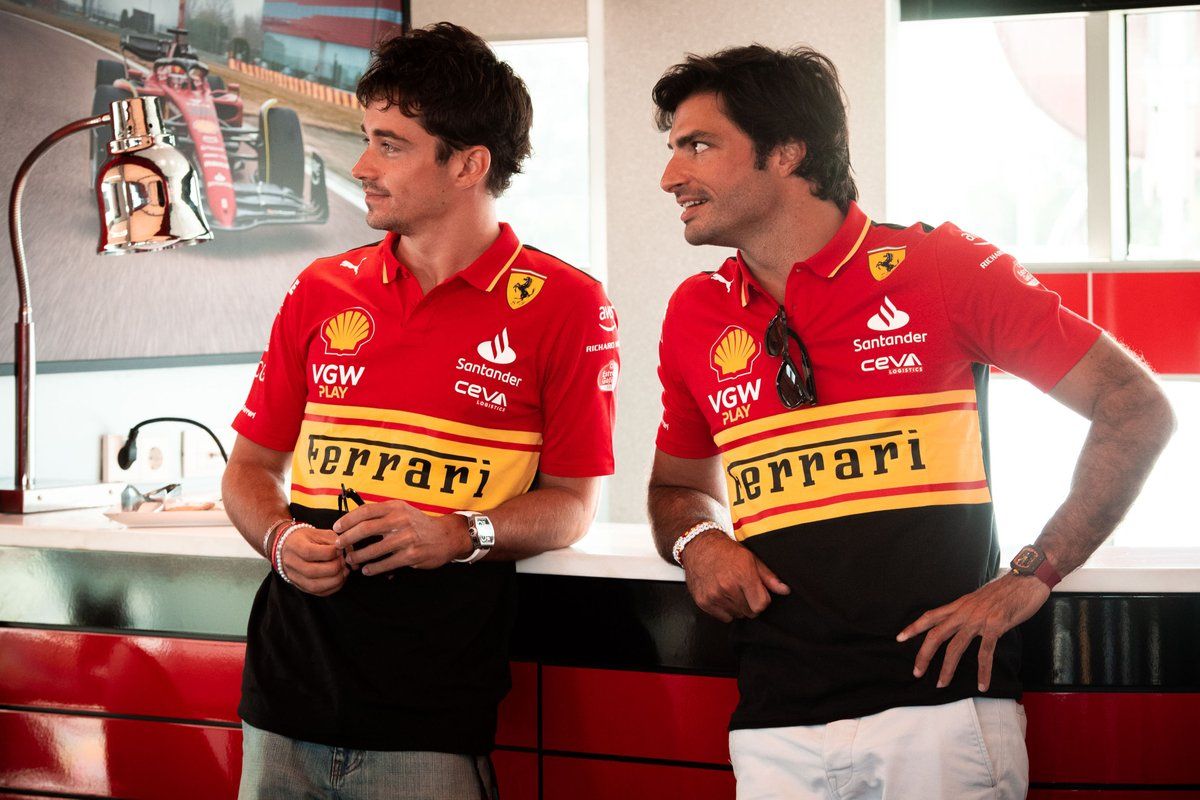 Charles Leclerc and Carlos Sainz in red and black Ferrari team polo shirts, leaning on a counter.