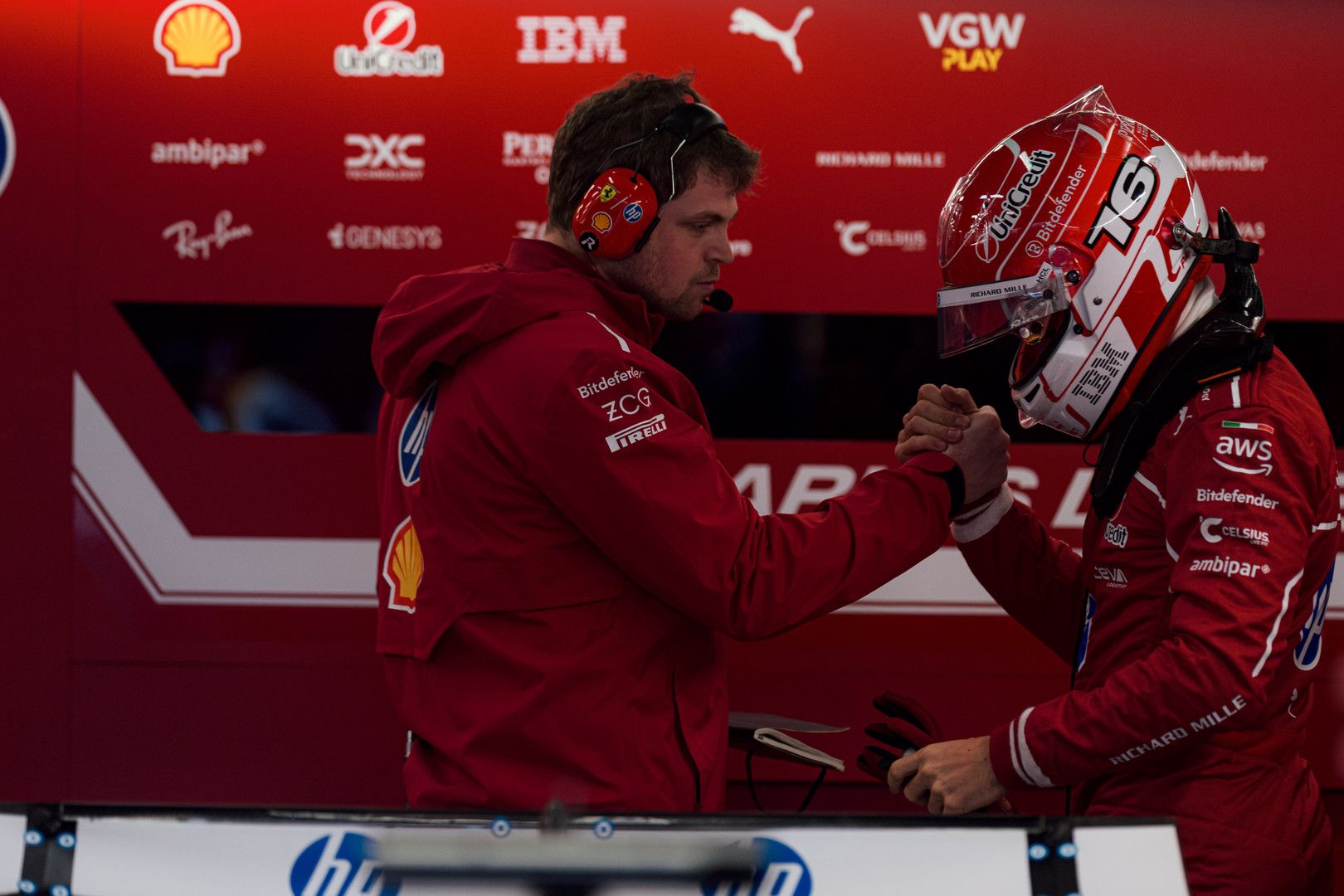 A racing driver in a red suit and helmet shakes hands with a team member in a red garage.