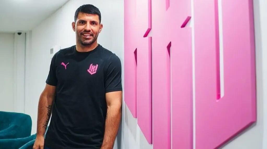 Sergio Agüero smiles, wearing a black shirt, leaning against a white wall with large pink 3D art.