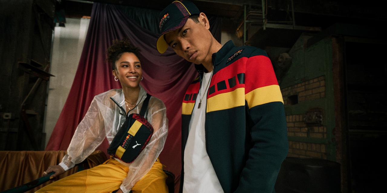 A smiling woman with a red and yellow fanny pack, and a man in a dark green, red, and yellow striped jacket and cap.