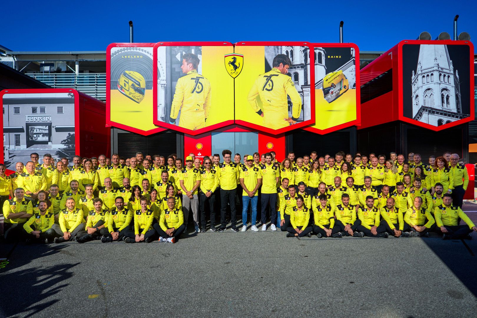 A large group of people in yellow shirts poses in front of a red structure adorned with Ferrari branding.