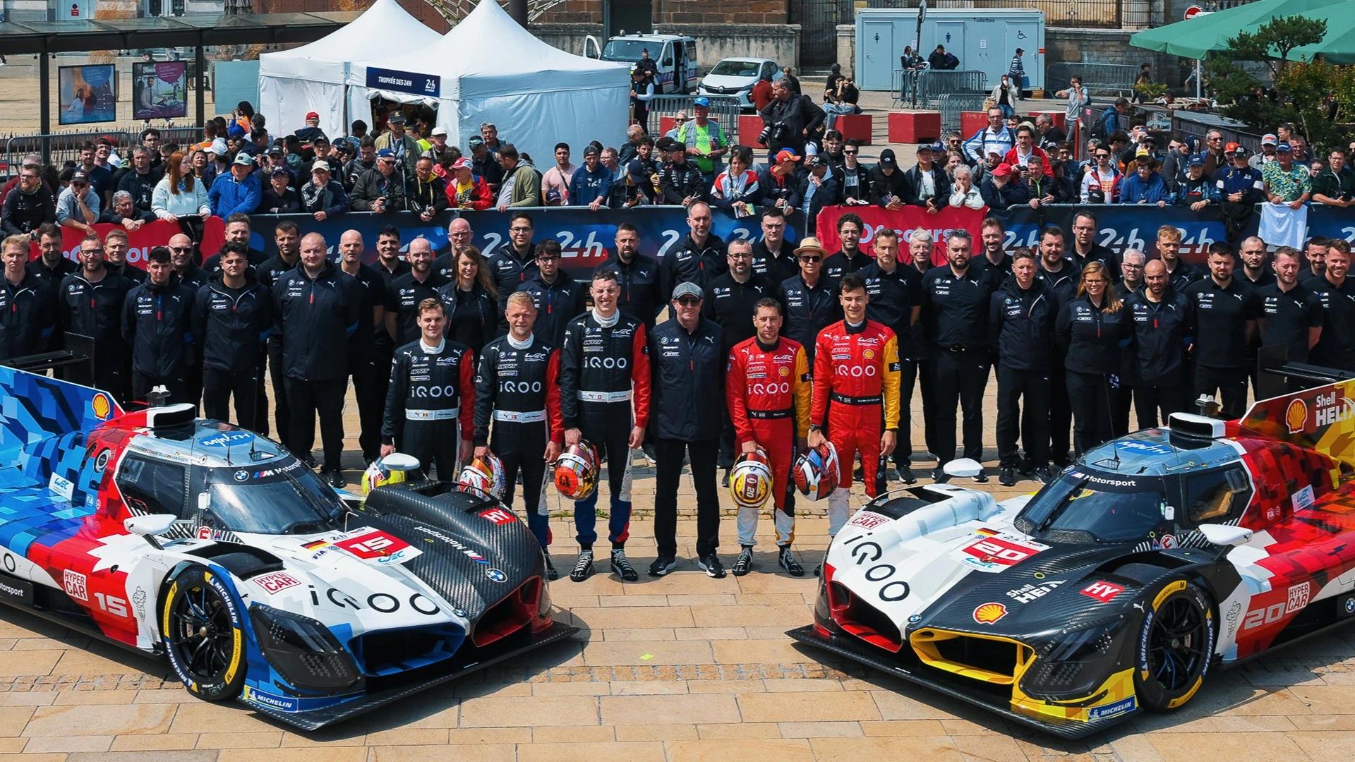 Two distinctively liveried prototype race cars, #15 and #20, flanked by their drivers and a large team, posing outdoors.