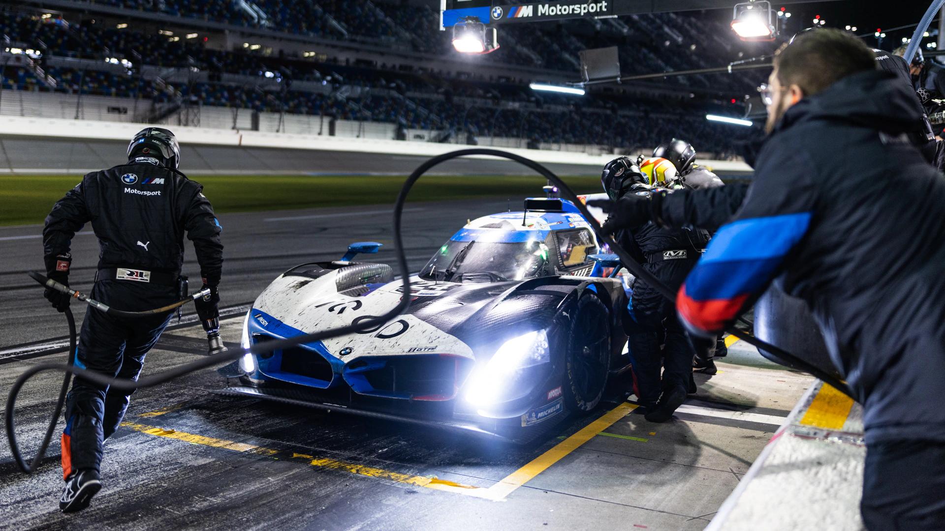 BMW M Motorsport race car with bright headlights during a night pit stop, surrounded by pit crew.