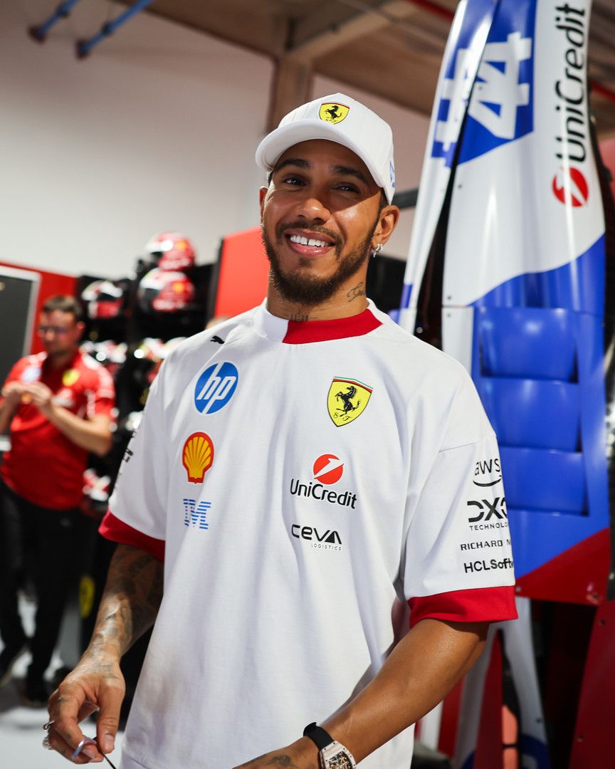 Lewis Hamilton smiles, wearing a white cap with a Ferrari logo and a white sponsored shirt.