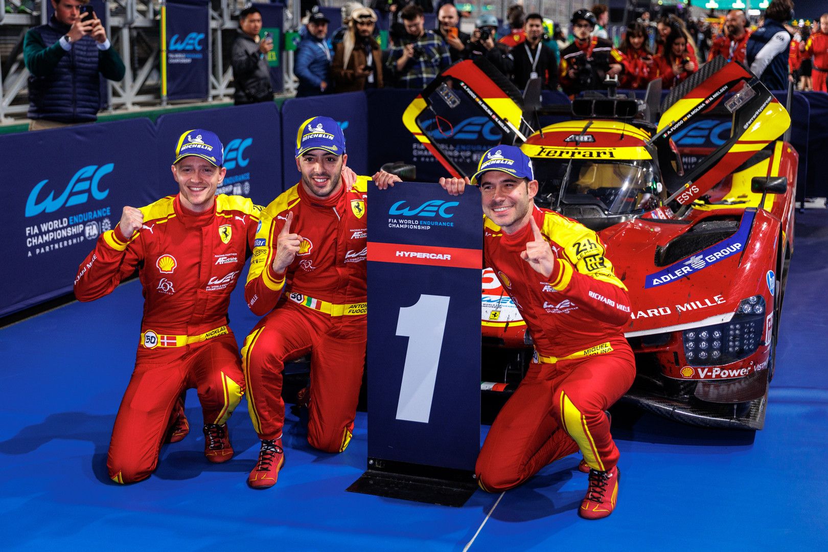 Three Ferrari WEC drivers celebrate with a "1" sign in front of their red hypercar.