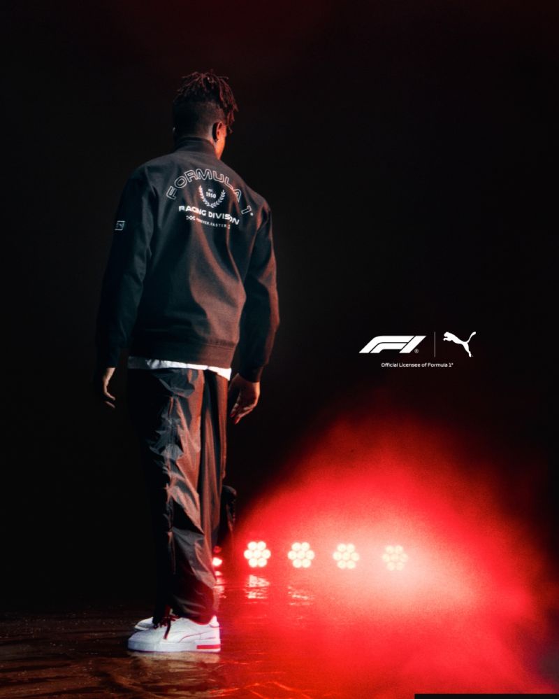 A person wears a black "Formula 1 Racing Division" jacket and white Puma sneakers in red stage lighting, with Formula 1 and Puma logos visible.