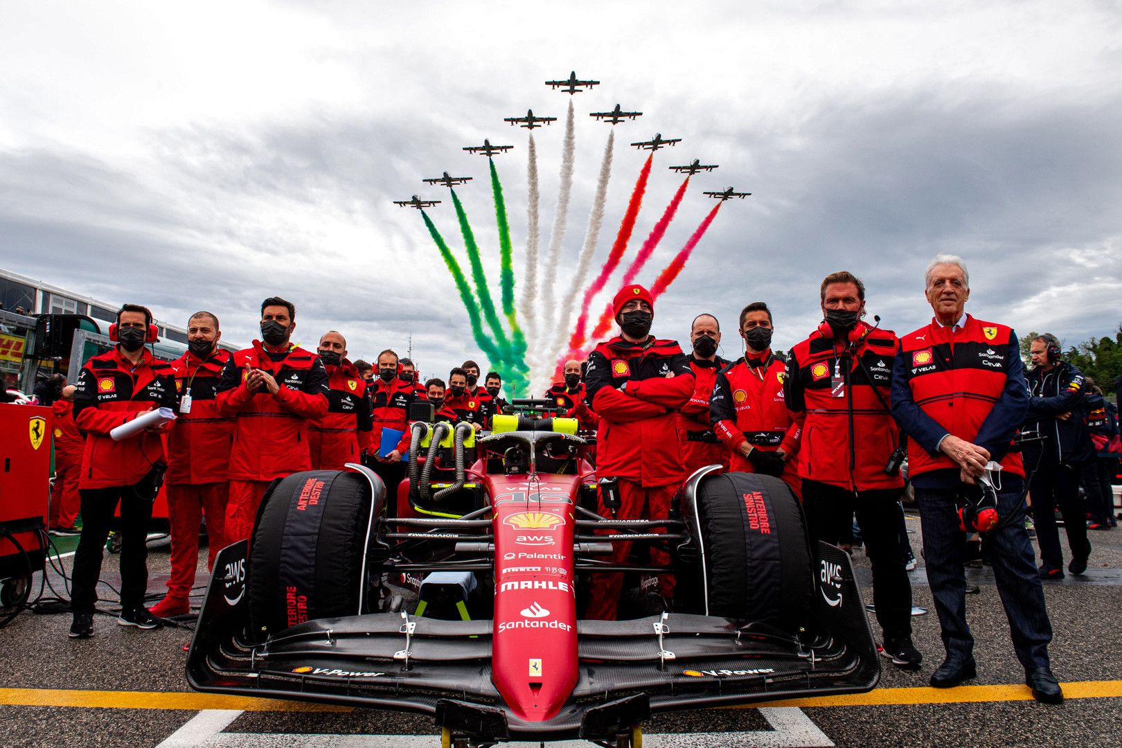 A Ferrari F1 car and team members in red stand on a track as jets fly overhead, trailing green, white, and red smoke.