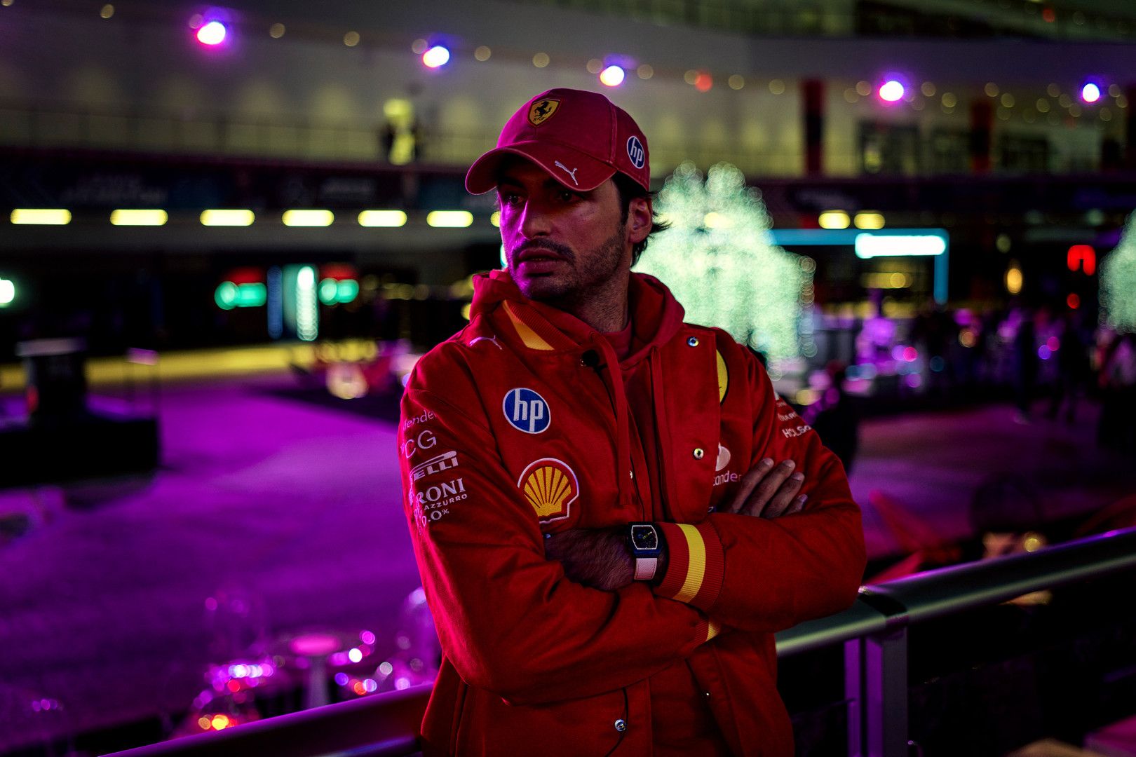 Carlos Sainz Jr. in a red Ferrari team jacket and cap, with arms crossed, against a dark, purple-lit background.