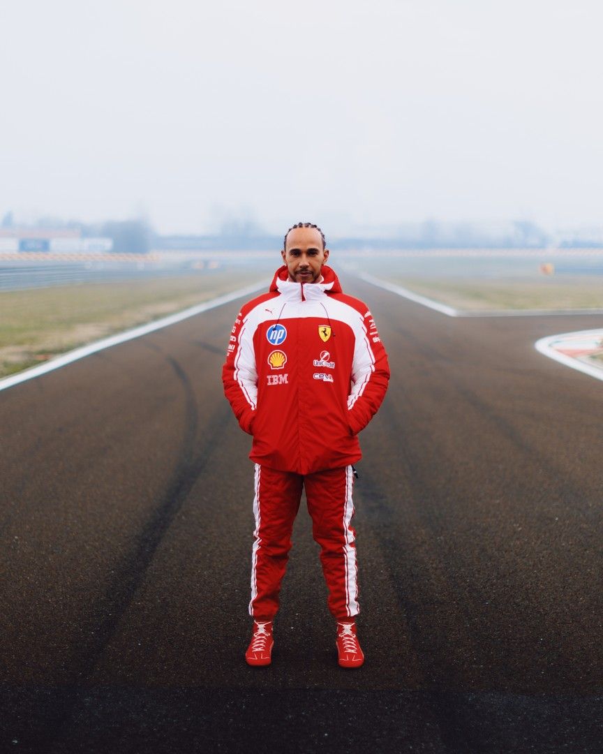 Lewis Hamilton in a red and white Ferrari tracksuit stands on a foggy race track.