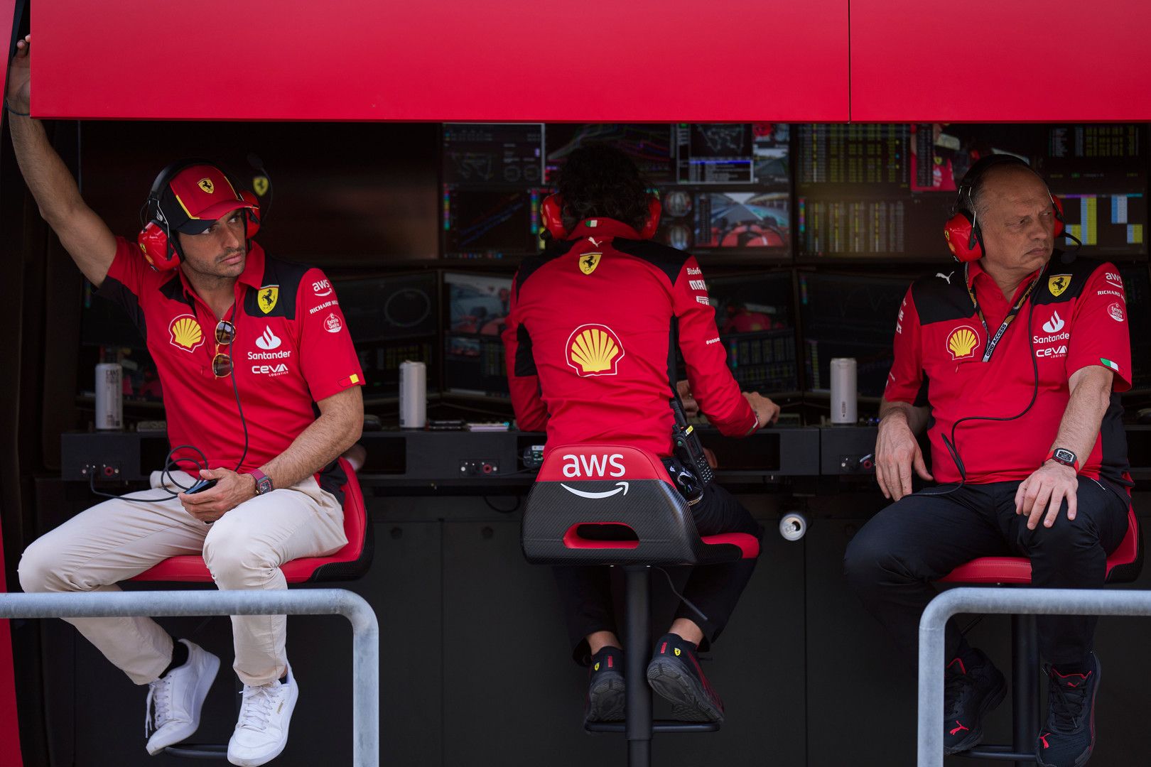 Three Ferrari team members in red shirts and headphones sit in a pit garage, looking at monitors.