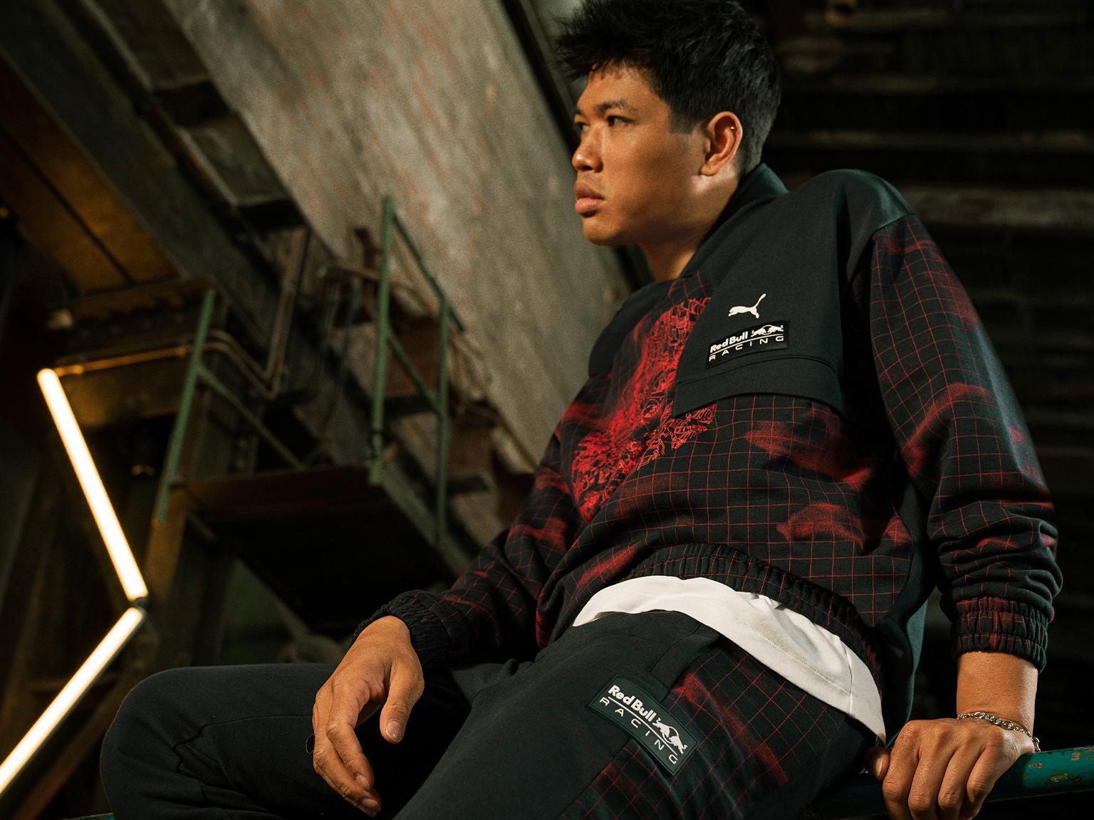 A man in a PUMA x Red Bull Racing tracksuit sitting and looking right in an industrial setting.
