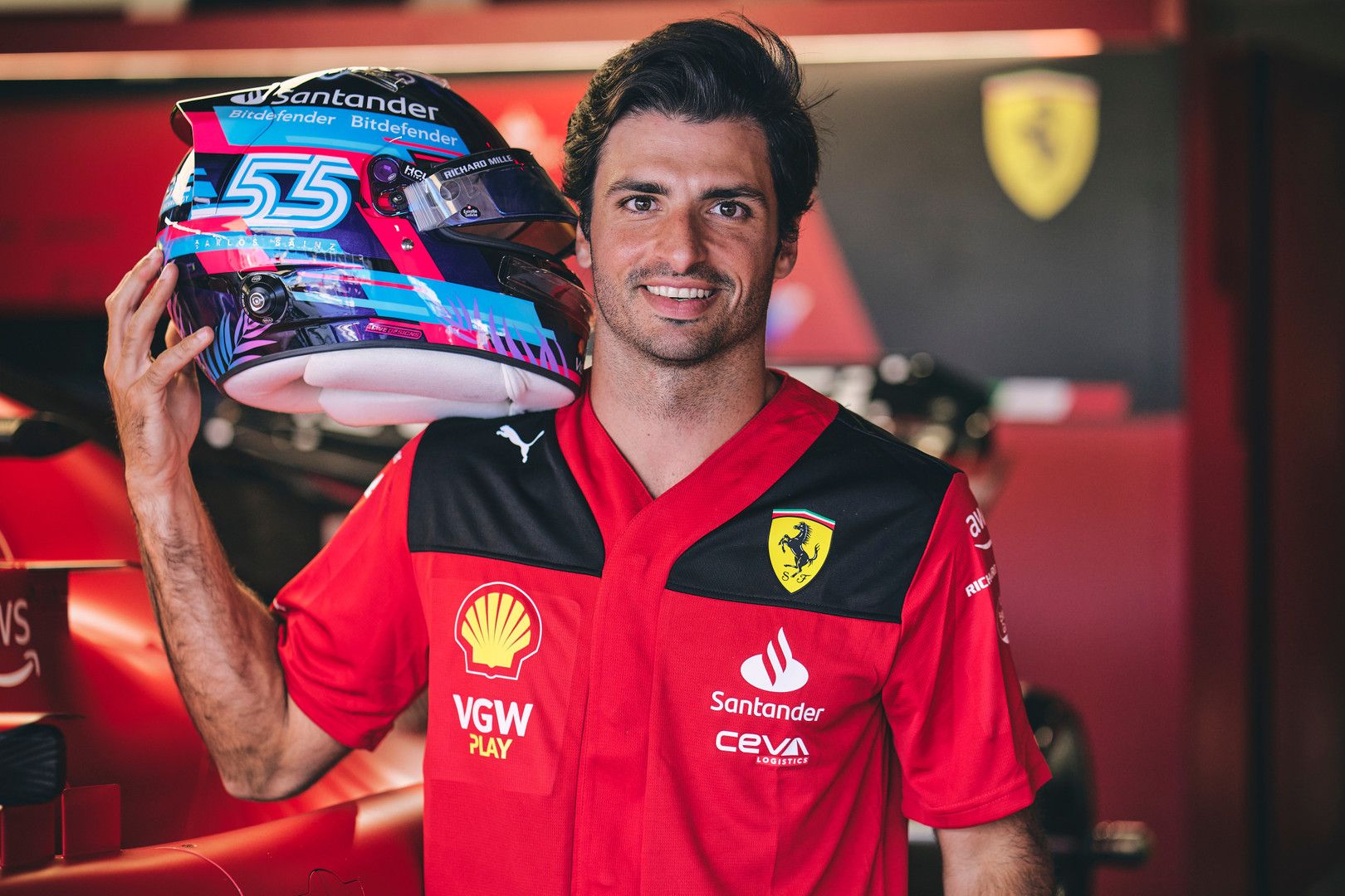 Carlos Sainz smiles, holding his vibrant racing helmet and wearing a red Ferrari team shirt.