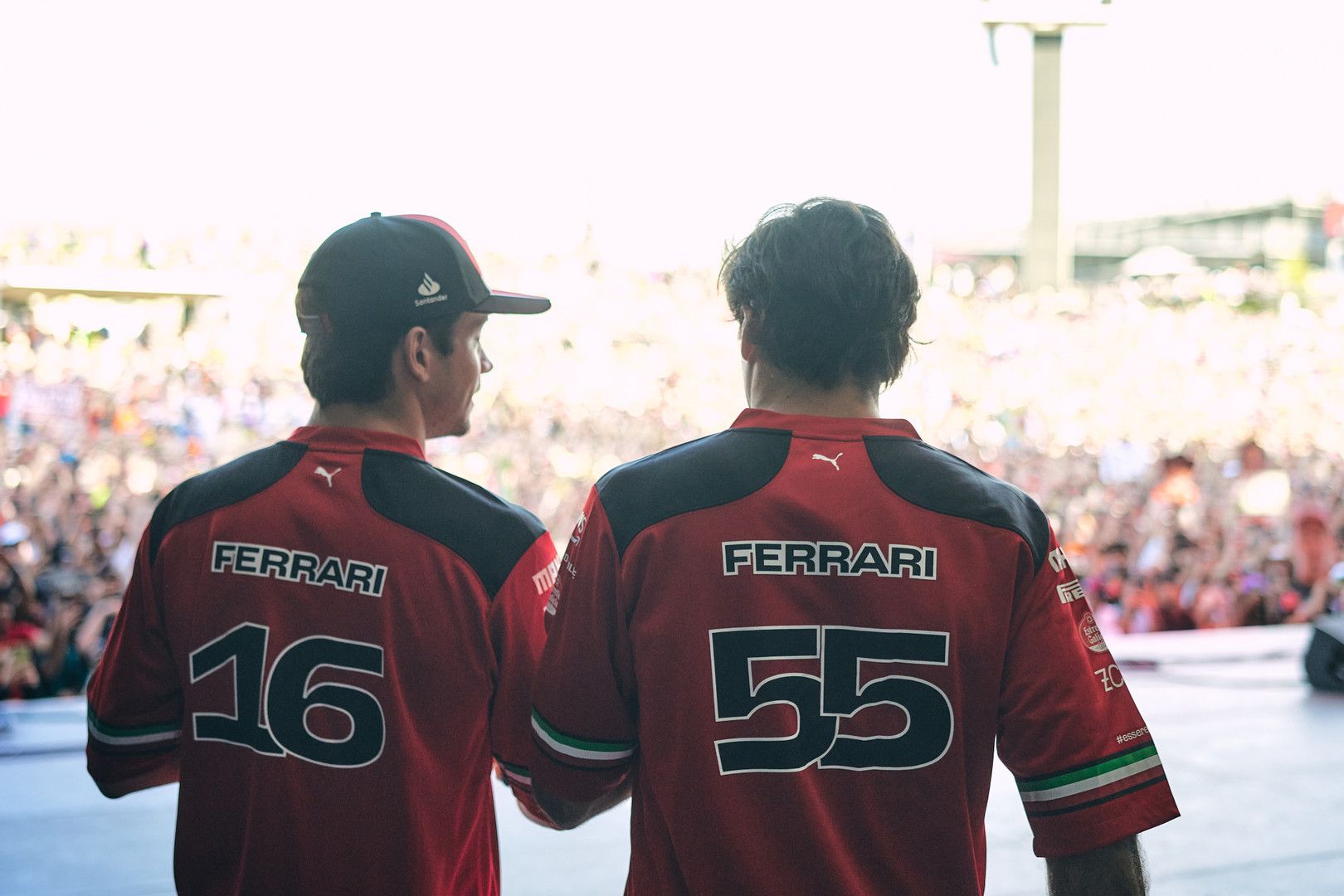 Two people in red Ferrari shirts with numbers 16 and 55 on their backs face a large crowd.