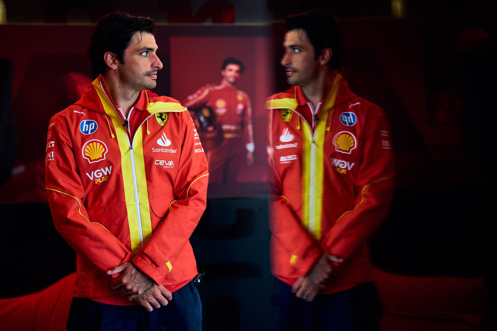 Carlos Sainz Jr. in a red Ferrari jacket, with his reflection and a blurred image of him in a racing suit in the background.