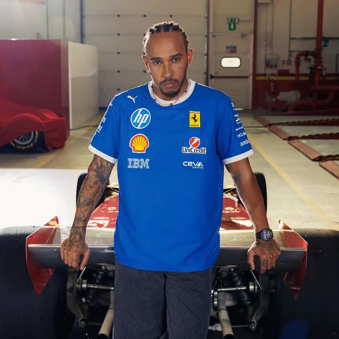 Lewis Hamilton in a blue Ferrari F1 team shirt standing in a garage with a race car.
