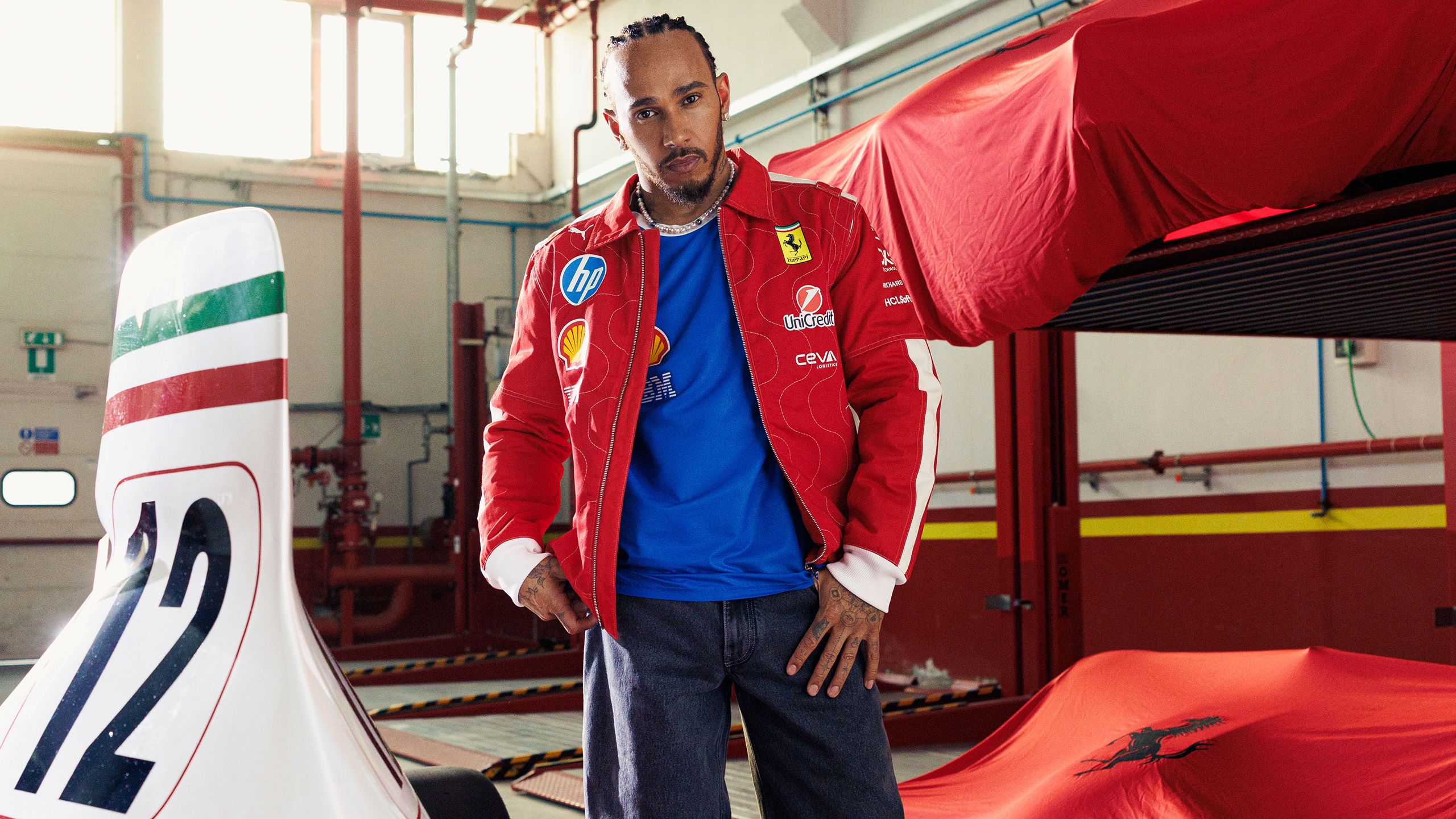 Lewis Hamilton in a red Ferrari jacket stands in a garage next to a white race car with the number 12.