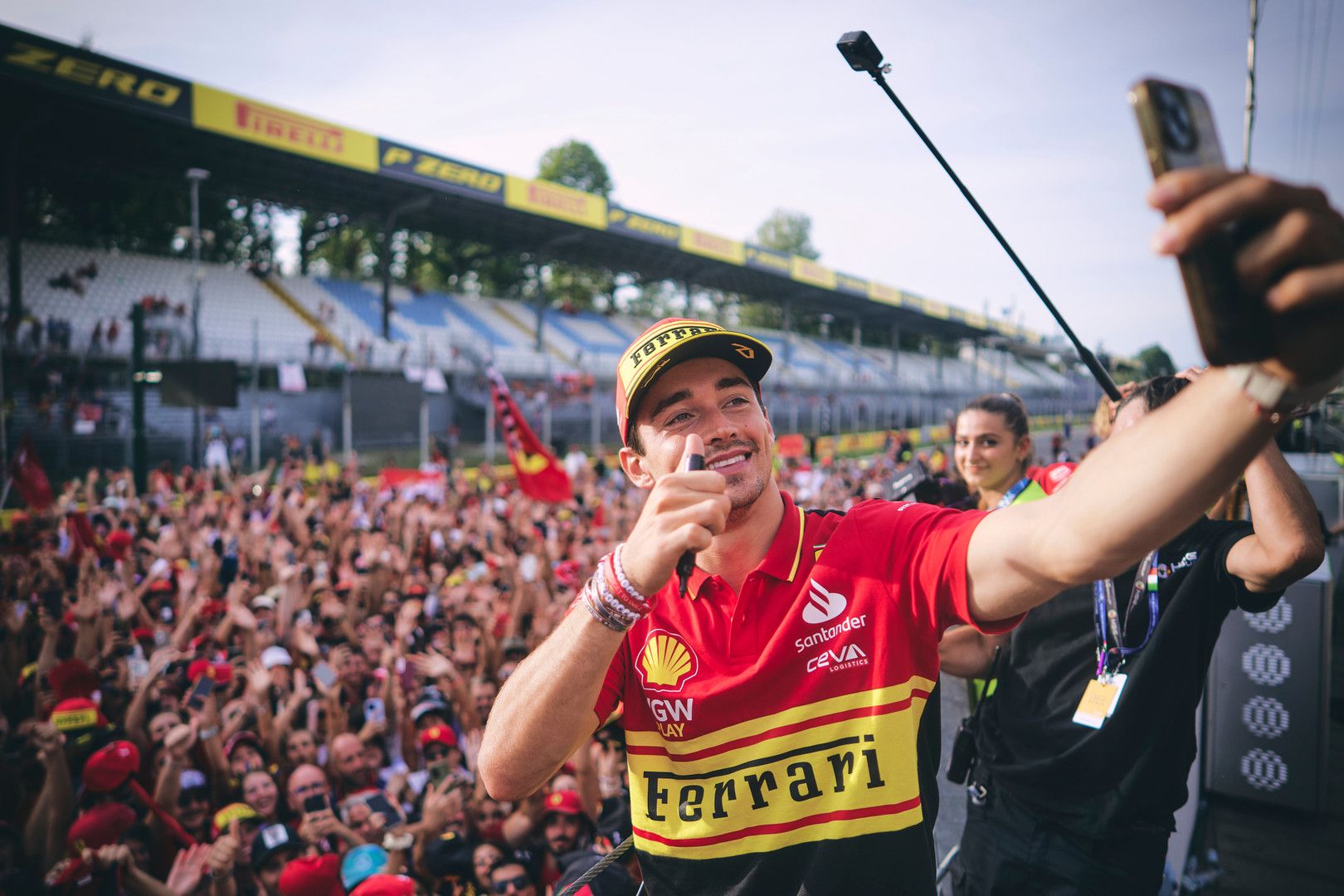 Charles Leclerc smiles and gives a thumbs-up while taking a selfie with a cheering crowd at a racetrack.