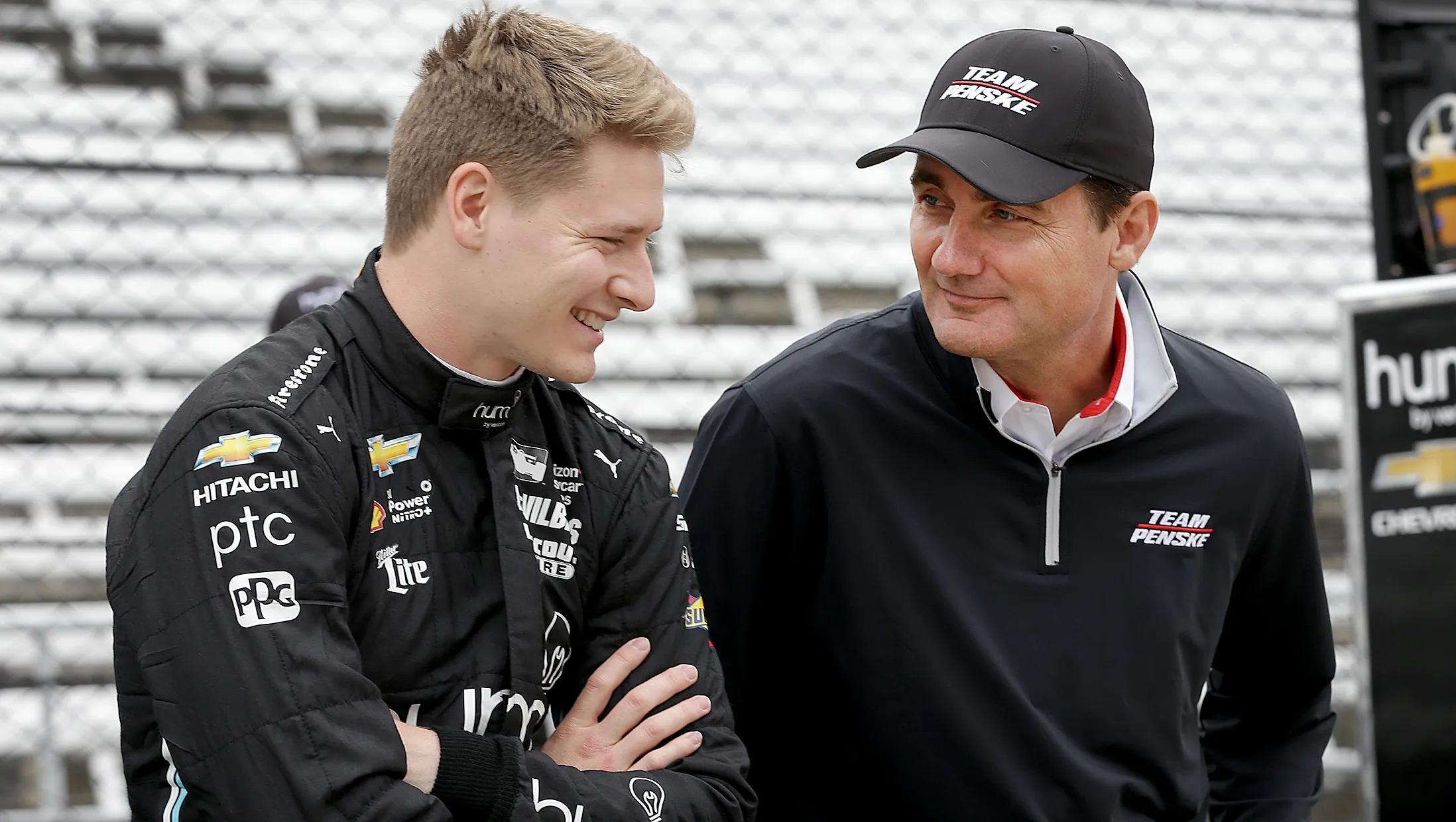A smiling race car driver in a black suit stands next to a man in a Team Penske cap and pullover at a racetrack.