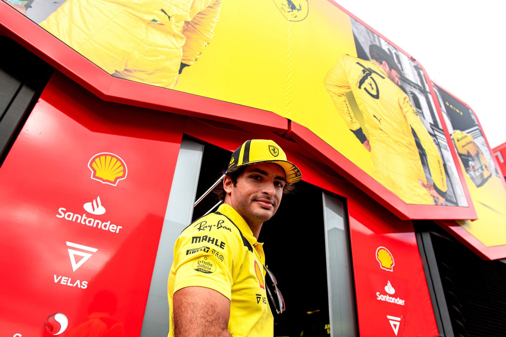 Carlos Sainz in a yellow Ferrari team polo shirt and cap, standing in front of a red team garage with sponsor logos and large yellow banners.