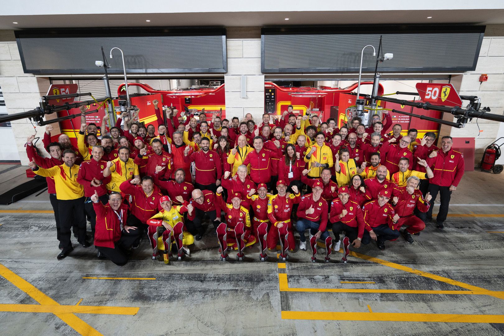 A large group of Ferrari racing team members in red and yellow uniforms celebrate in a pit garage.