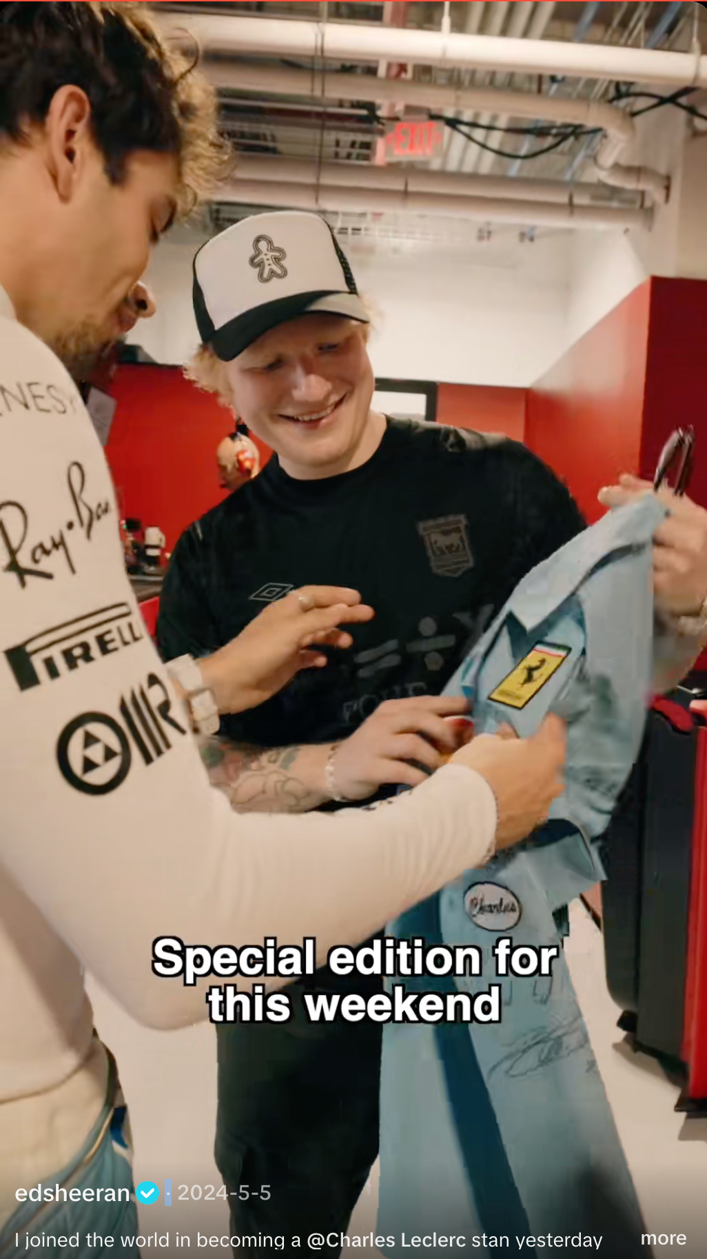 Ed Sheeran smiles, wearing a black t-shirt and gingerbread man hat, as a person in a white racing team shirt presents him with a light blue Charles Leclerc racing suit.