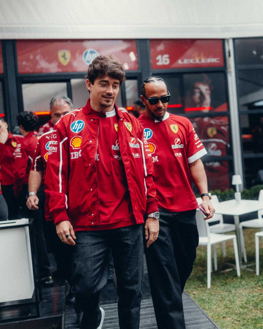 Charles Leclerc walks ahead of Lewis Hamilton in a paddock, both wearing red Ferrari team attire, with Hamilton wearing sunglasses.