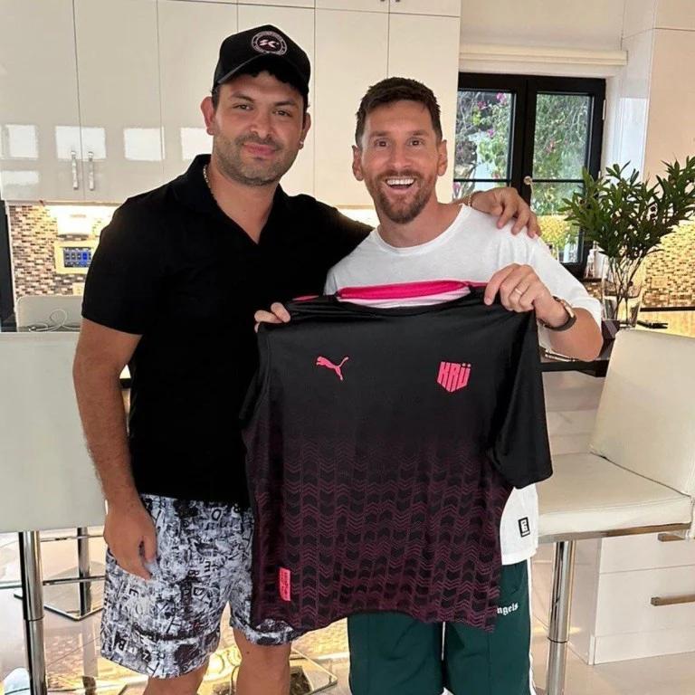 Lionel Messi and another man smiling and holding a black and pink sports jersey.