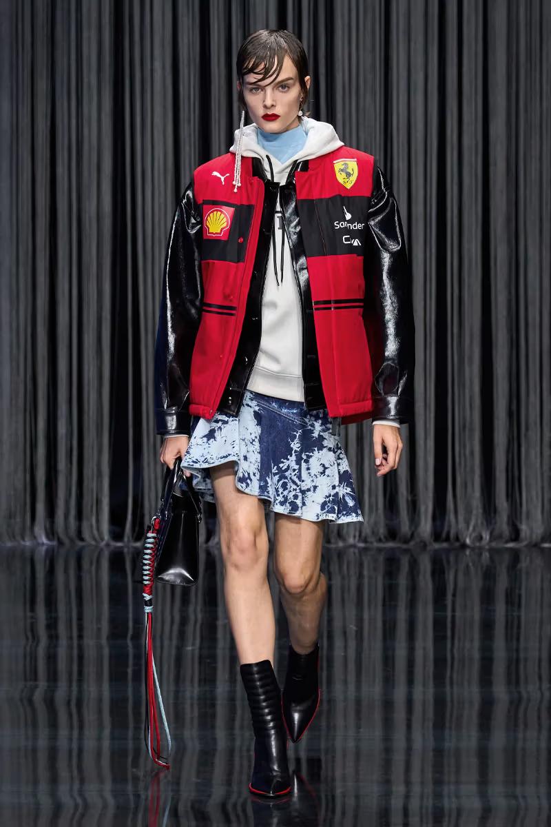 Androgynous model with red lipstick in a red and black varsity jacket, white hoodie, tie-dye denim mini-skirt, black boots, and a black bag on a runway.