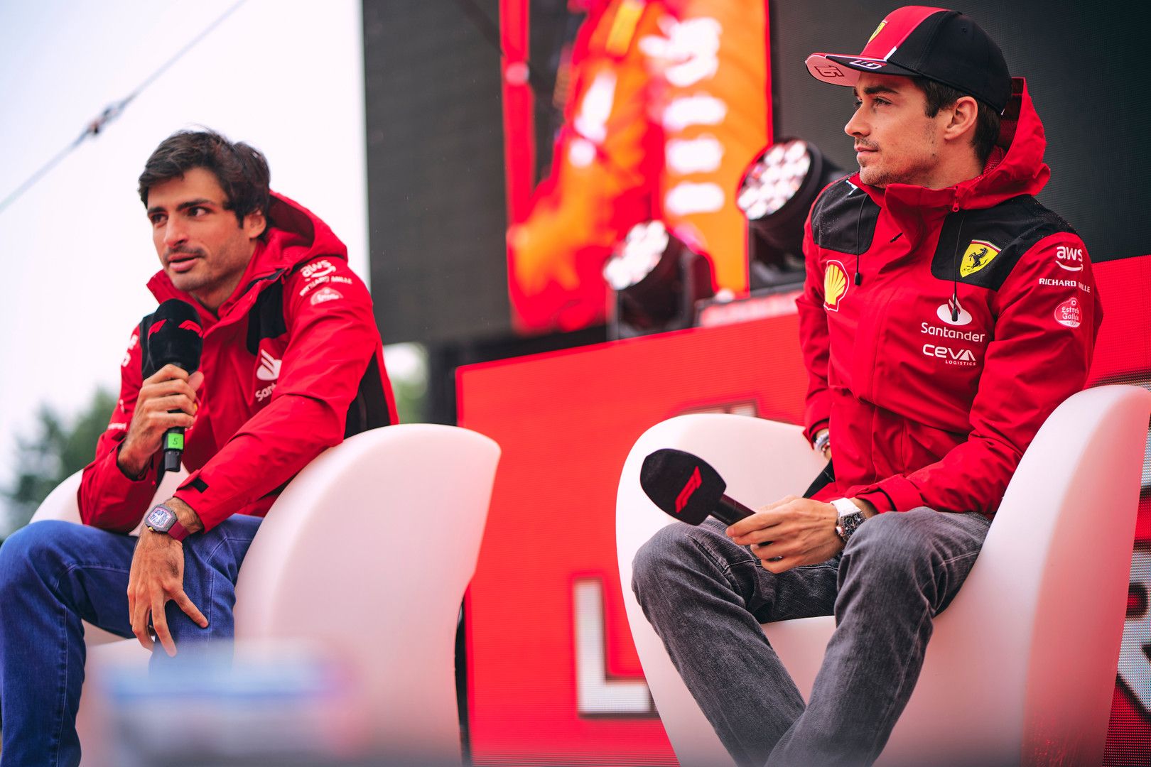 Carlos Sainz and Charles Leclerc in red Ferrari jackets sit holding microphones.