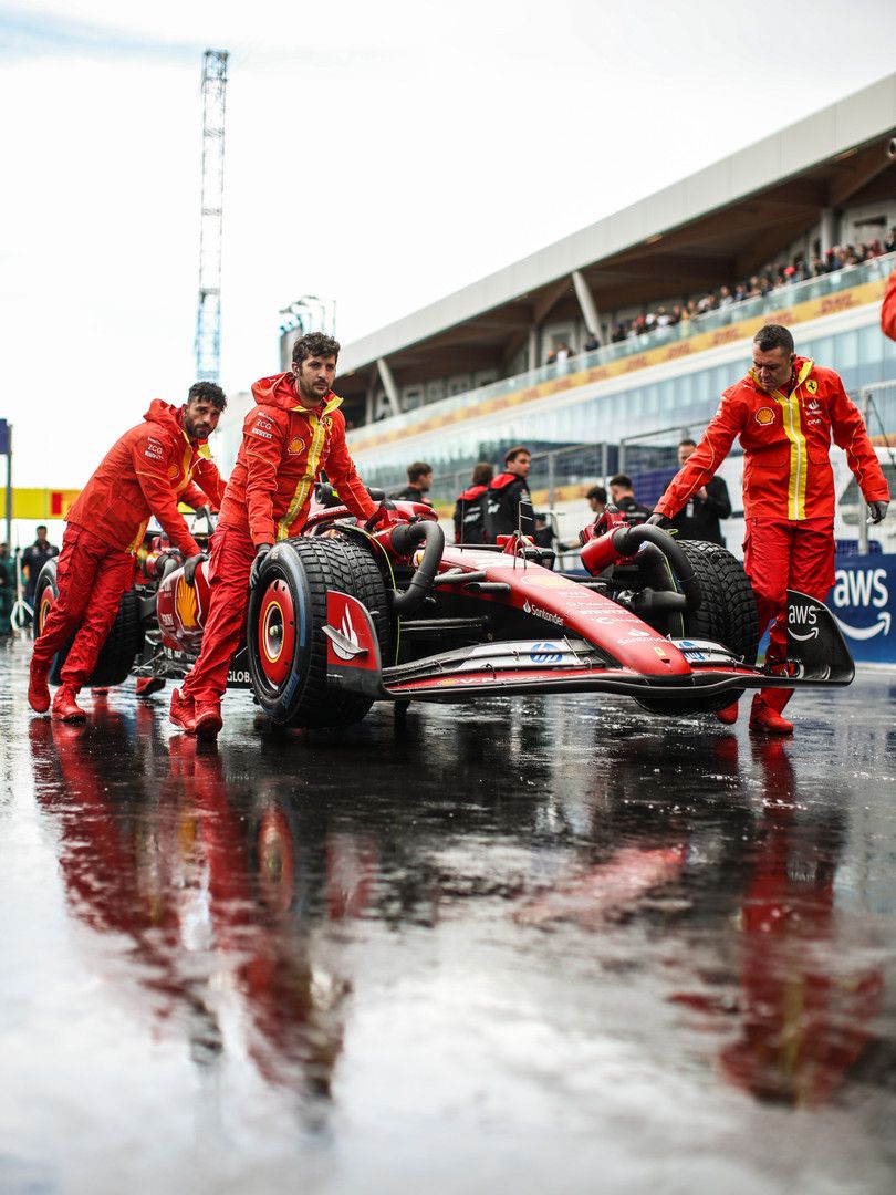 Ferrari team members push a red F1 car on a wet track.