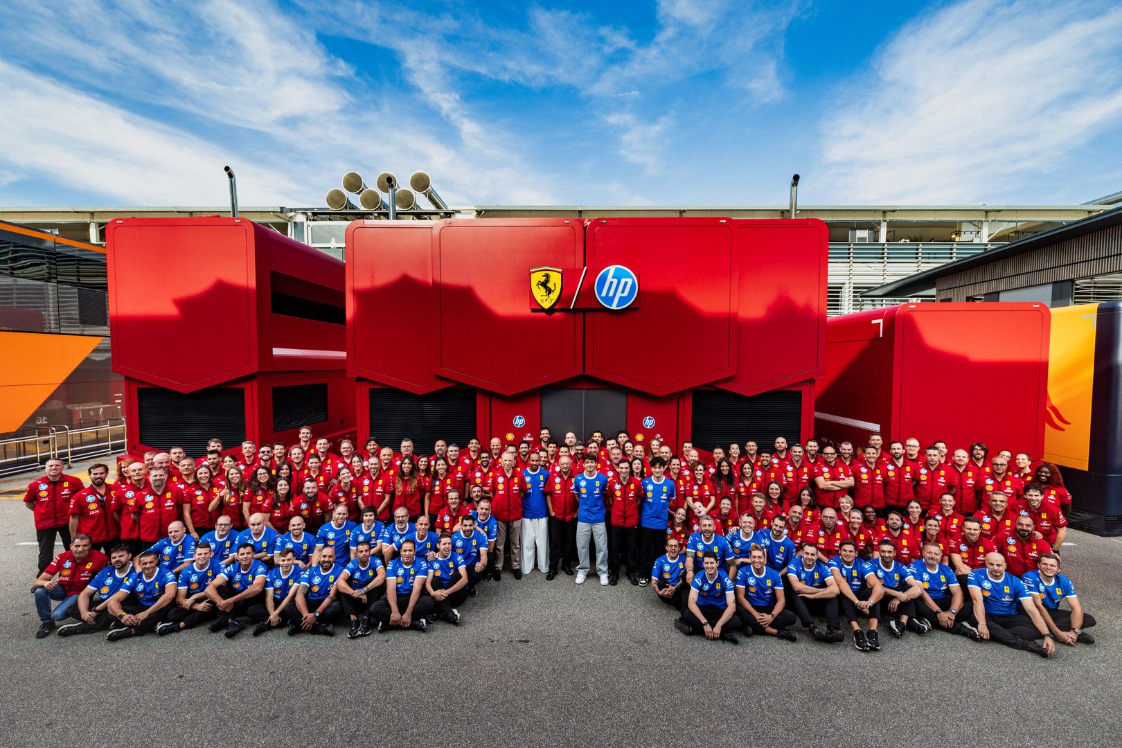 A large team in red and blue uniforms poses in front of a red building featuring Ferrari and HP logos.