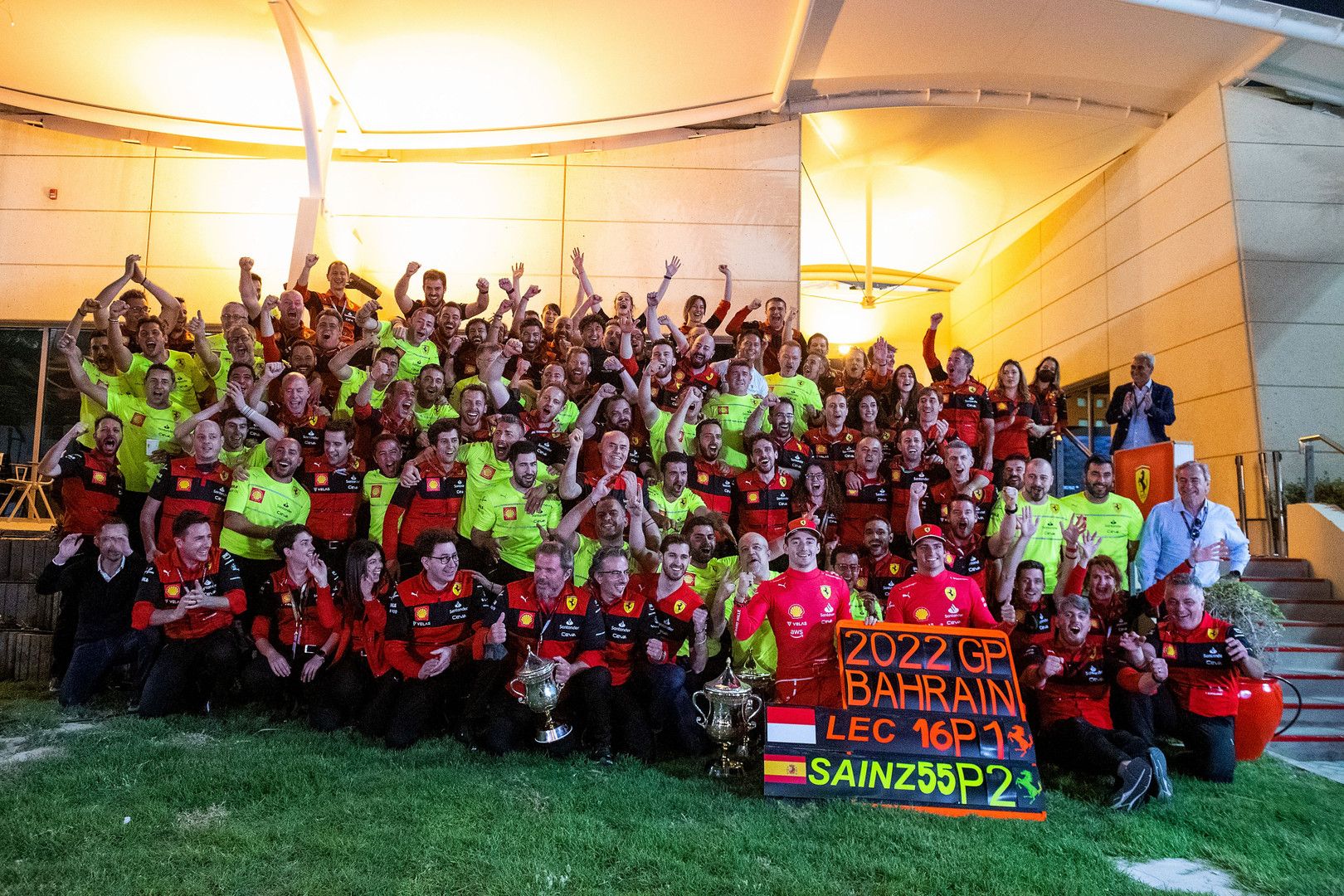The Ferrari F1 team celebrates their 1st and 2nd place victory at the 2022 Bahrain Grand Prix, posing with trophies and a celebratory sign.
