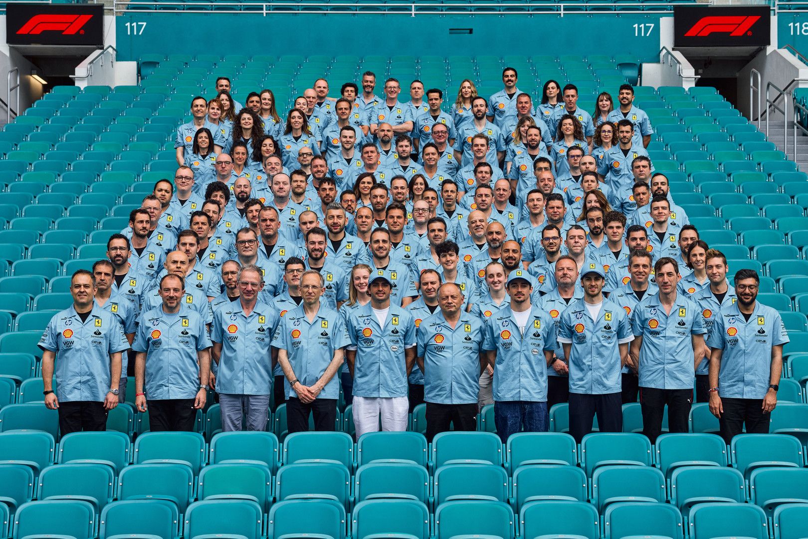 A large group of people in light blue shirts with Shell logos pose in a stadium's turquoise seats.