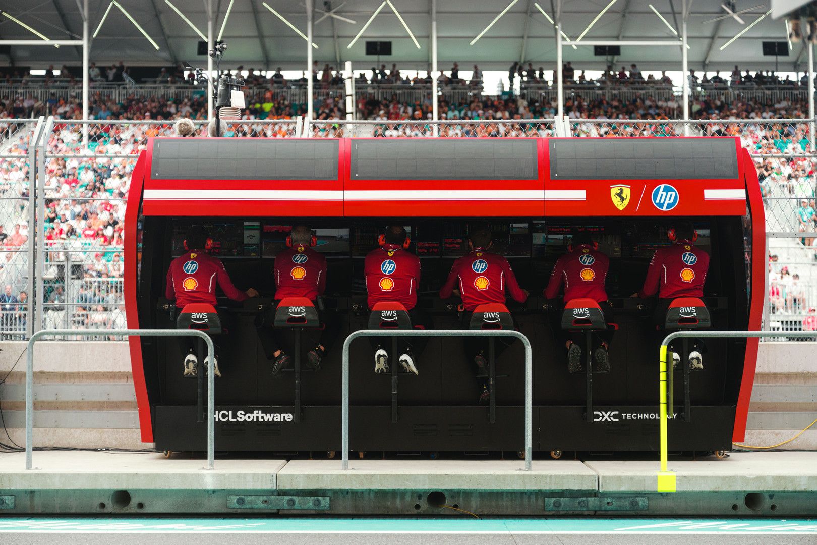 Six F1 Ferrari team members in red uniforms sit in a pit wall booth, with grandstands and spectators in the background.